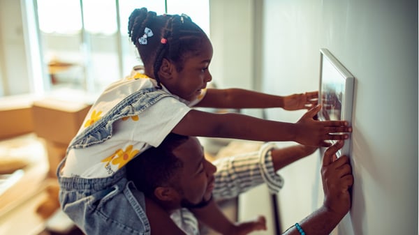 A child sits on an adult’s shoulders as they hang a picture frame on the wall of their new home.