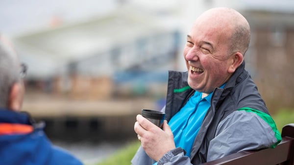 Two people sit on a wooden bench in an outdoor setting. One wears a blue shirt and grey jacket with green accents, holding a black coffee cup.