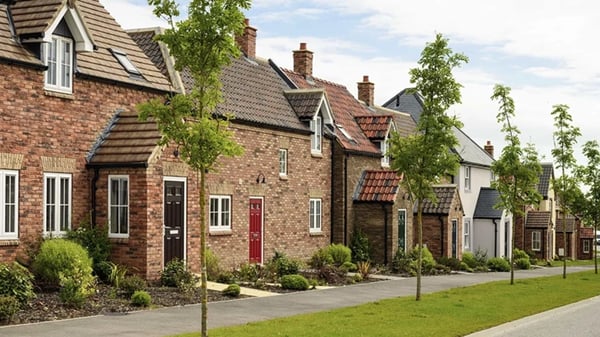 Street view of a suburban housing development; a row of traditional-style brick homes with pitched roofs and varied door colours. Each home has a landscaped front garden, and young trees line a pedestrian pathway.