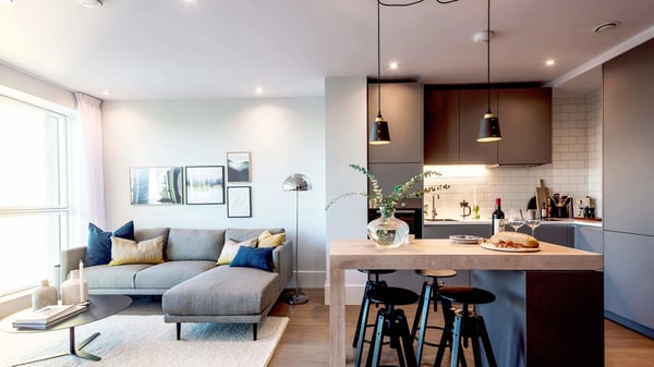 Modern open-plan living space with a light grey sectional sofa and framed artwork on the wall. The space transitions into a sleek kitchen with dark grey cabinetry, white subway tile backsplash, and a wooden island with three black bar stools. Black pendant lights hang above the island, which is set with a glass vase, bottle of wine, and loaf of bread. A large window on the left brings in natural light.