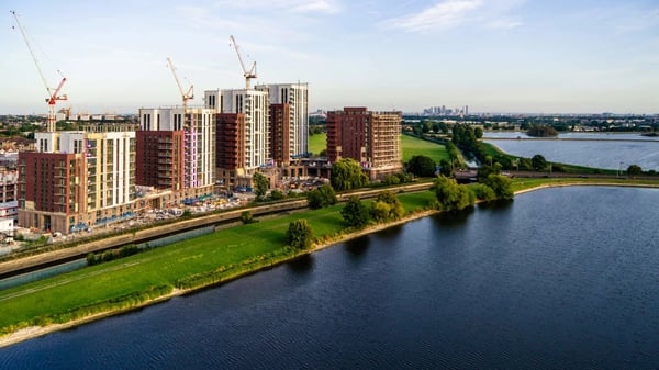 Aerial view of the New Acres scheme in Wandsworth, London. The image captures a vibrant new community taking shape, with contemporary mid-rise residential buildings under construction and cranes positioned across the site. The scheme is set alongside expansive water and green spaces, creating a balance of urban living and natural surroundings. In the distance, the London skyline is visible under a clear blue sky.