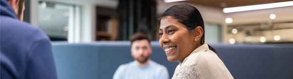 Two colleagues are seated around a table in a well-lit room with wooden ceiling panels. One wears a white and blue speckled jumper, another a navy blue hoodie.,