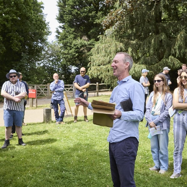 A group of people gathered outdoors on a grassy area with trees in the background, listening to a person holding papers and gesturing while speaking.