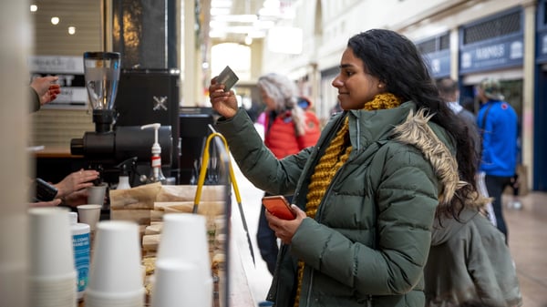 A mid adult woman on a sunny winters day. She is wearing casual, winter clothing and accessories. She is paying for a coffee with her credit card at a small coffee shop in a shopping arcade.