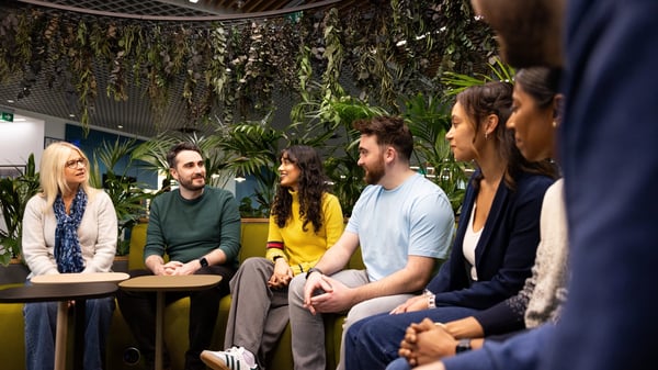 A group of people are seated in a semi-circle on green couches in Legal & General’s Cardiff office. The space is adorned with plants hanging from the ceiling.
