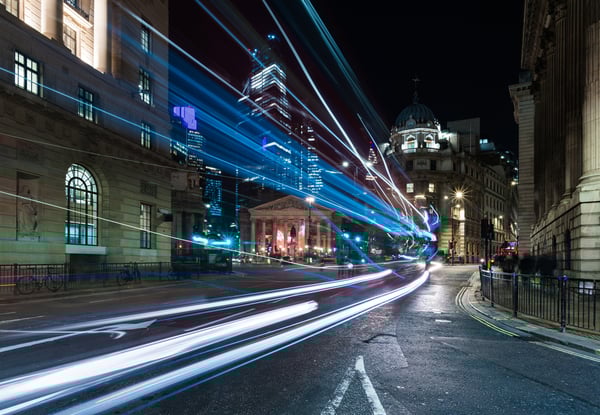 Night view of the Bank Junction in London, UK, with illuminated buildings and skyscrapers in the background. Bright streetlights create starburst effects, and light trails from passing vehicles flow across the foreground.